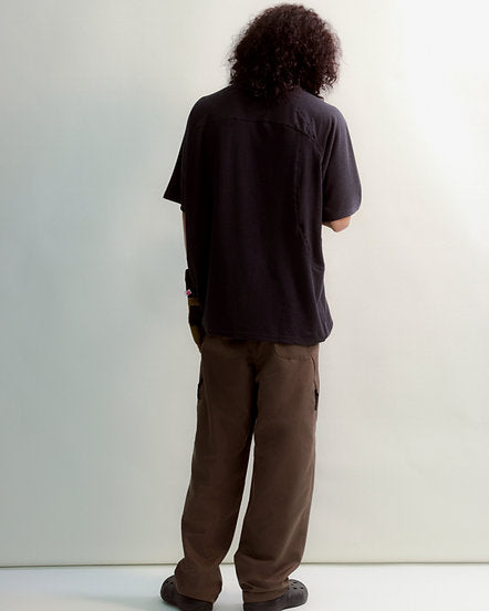 Person wearing a black t-shirt and brown pants standing against a white wall.