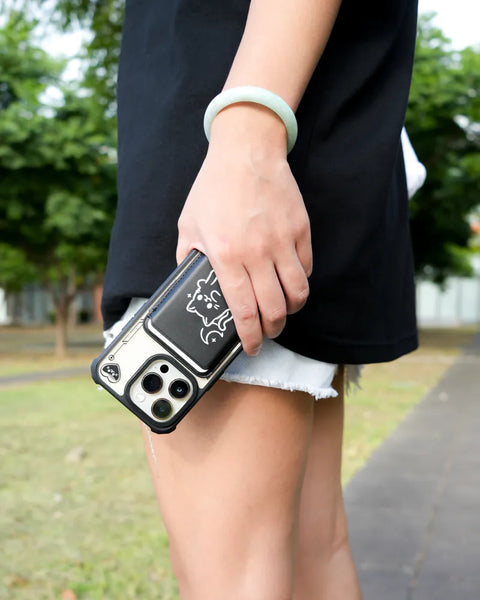Person holding a phone with a black case featuring a white design, outdoors.