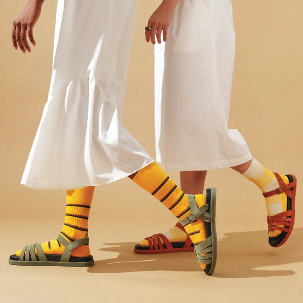 Two people wearing white pants and colorful sandals with yellow socks on a beige background
