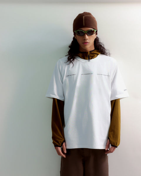 Person wearing a white t-shirt with long sleeves in brown and green, sunglasses, and a cap on a plain background