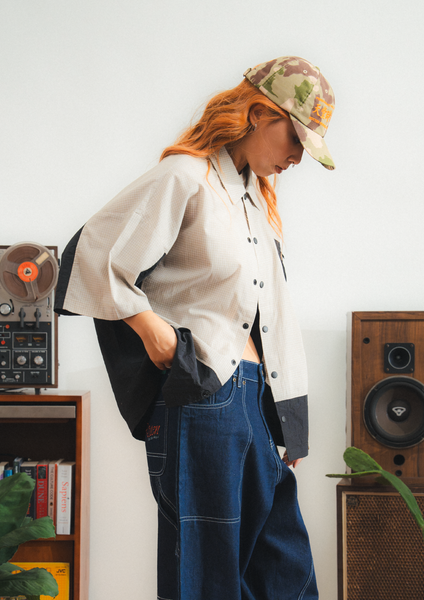 Person wearing a white shirt and blue jeans with a camouflage cap, standing in a room with speakers and books.