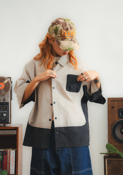 Person wearing a layered outfit with a camouflage cap in a room with vintage speakers and books.