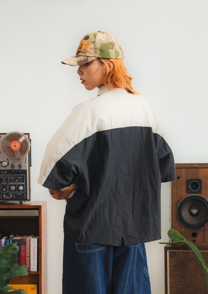 Person wearing a two-tone jacket and cap in a room with vintage speakers and records.