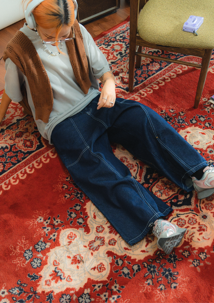 Person sitting on a patterned rug wearing a brown vest and blue jeans.