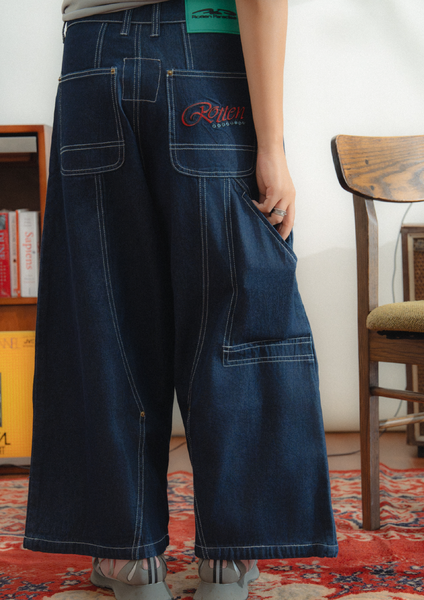 Person wearing dark blue wide-leg jeans with a brand logo, standing in a room with a chair and books.