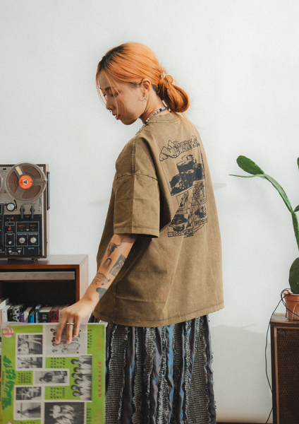 Person wearing a brown jacket with graphic designs, standing in a room with a plant and record player.