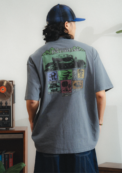 Person wearing a gray t-shirt with a colorful graphic on the back, standing in a room with a white wall and wooden shelf.