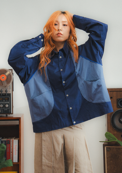 Person wearing a blue and navy color-blocked shirt with beige pants, standing in a room with vintage speakers and books.