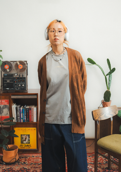 Person wearing a brown cardigan and headphones in a room with a vintage reel-to-reel tape recorder and books.
