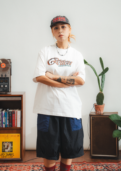 Person wearing a white t-shirt with text and dark shorts in a room with a plant and record player.