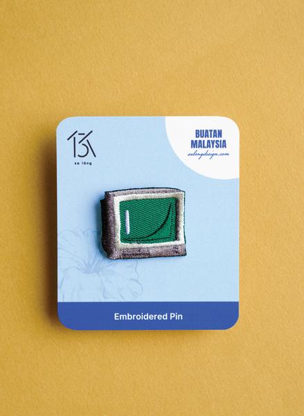 Embroidered pin on a blue card with 'Buatan Malaysia' branding against a mustard yellow background