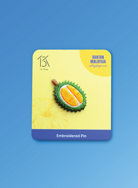 Embroidered pin of a durian fruit on a yellow card with 'Buatan Malaysia' branding against a blue background.