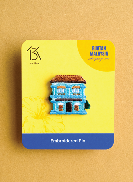 Embroidered pin of a building on a yellow card with 'Buatan Malaysia' branding.