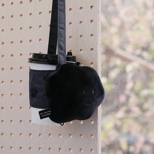 Black plush bag with a smiley face design attached to a coffee cup on a pegboard.