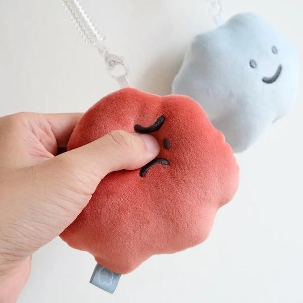 Plush toy resembling a peach with a sad face held by a hand, with another plush toy in the background.
